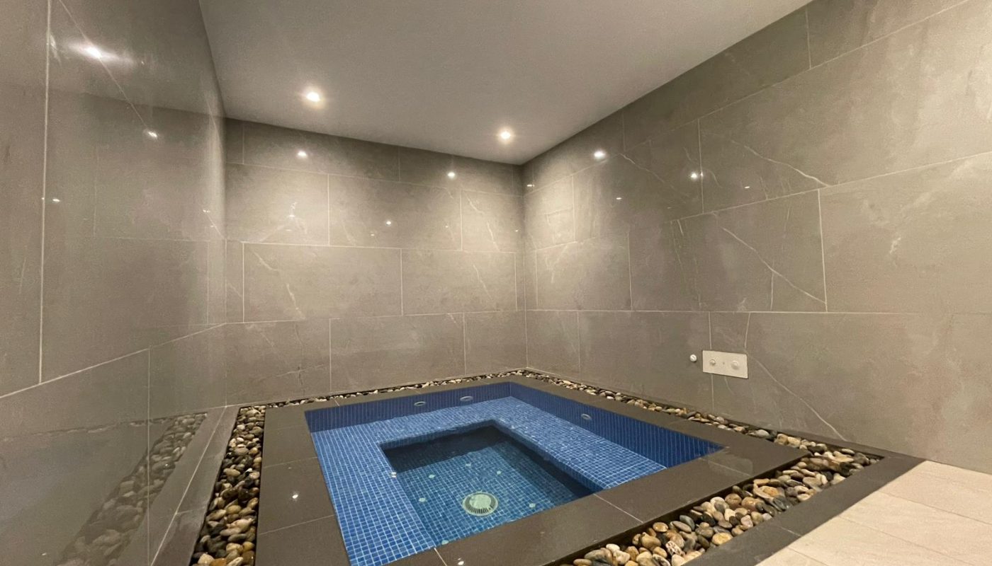 A small indoor square jacuzzi with blue tiles, surrounded by gray marble walls and decorative pebbles along the edges, under recessed ceiling lights.