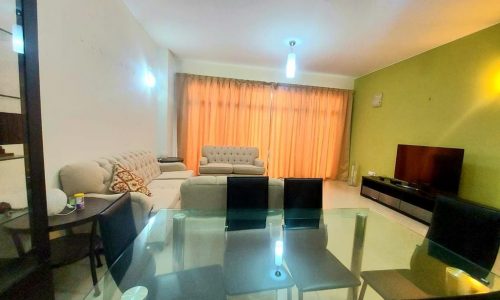 Spacious flat for sale in Amwaj with a living room featuring a gray sofa, matching loveseat, glass dining table with four chairs, and a television unit, illuminated by ceiling lights, with orange curtains.