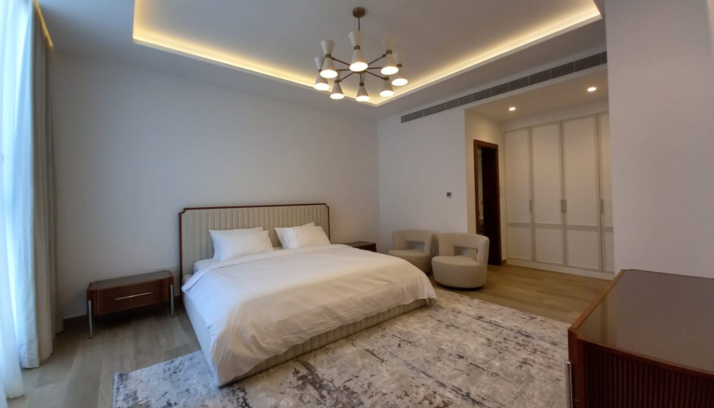 Modern bedroom with a double bed, two chairs, a large rug, wooden flooring, built-in wardrobe, and a ceiling light fixture. Natural light comes through a window with sheer curtains.