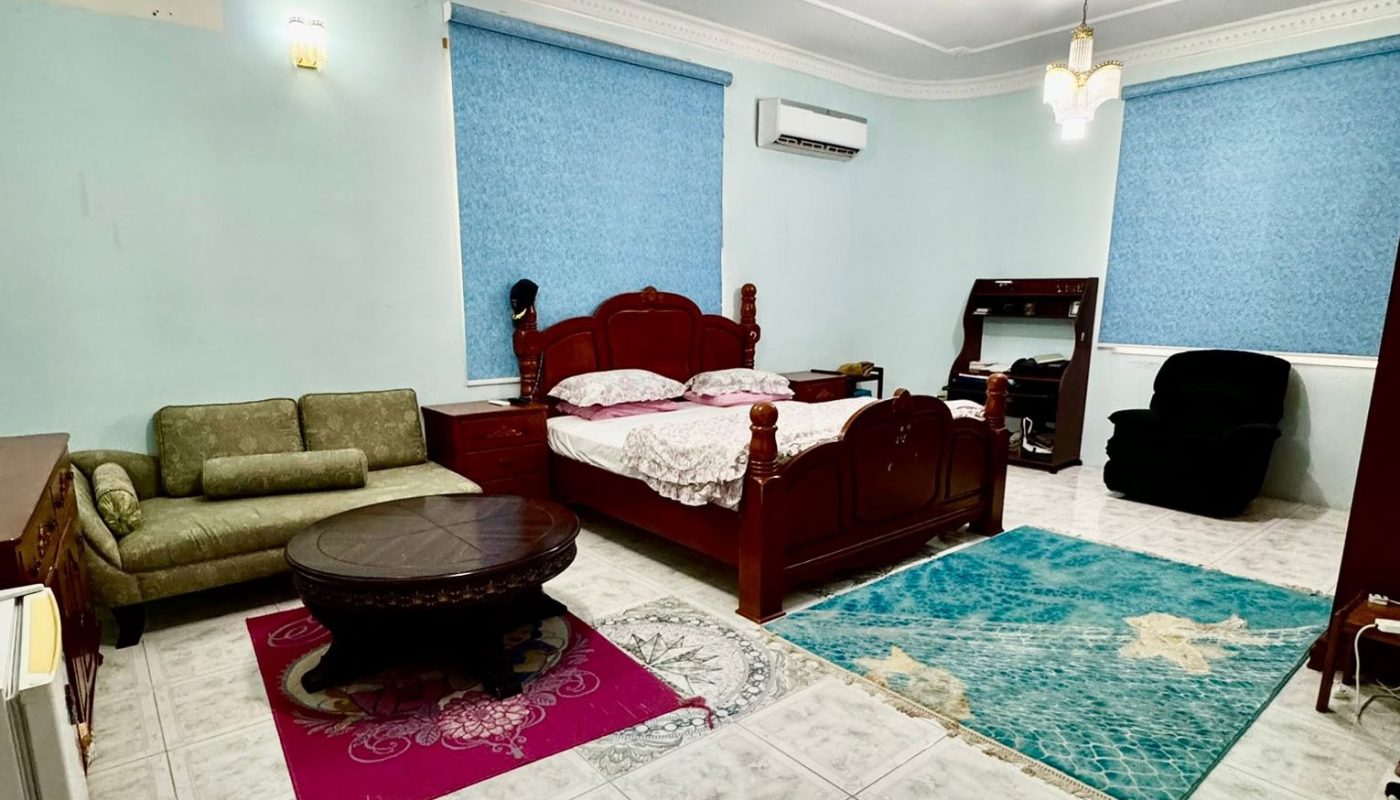 Spacious bedroom with a wooden bed, green sofa, round coffee table, desk, black chair, blue curtains, ceiling fan, and tiled floor.