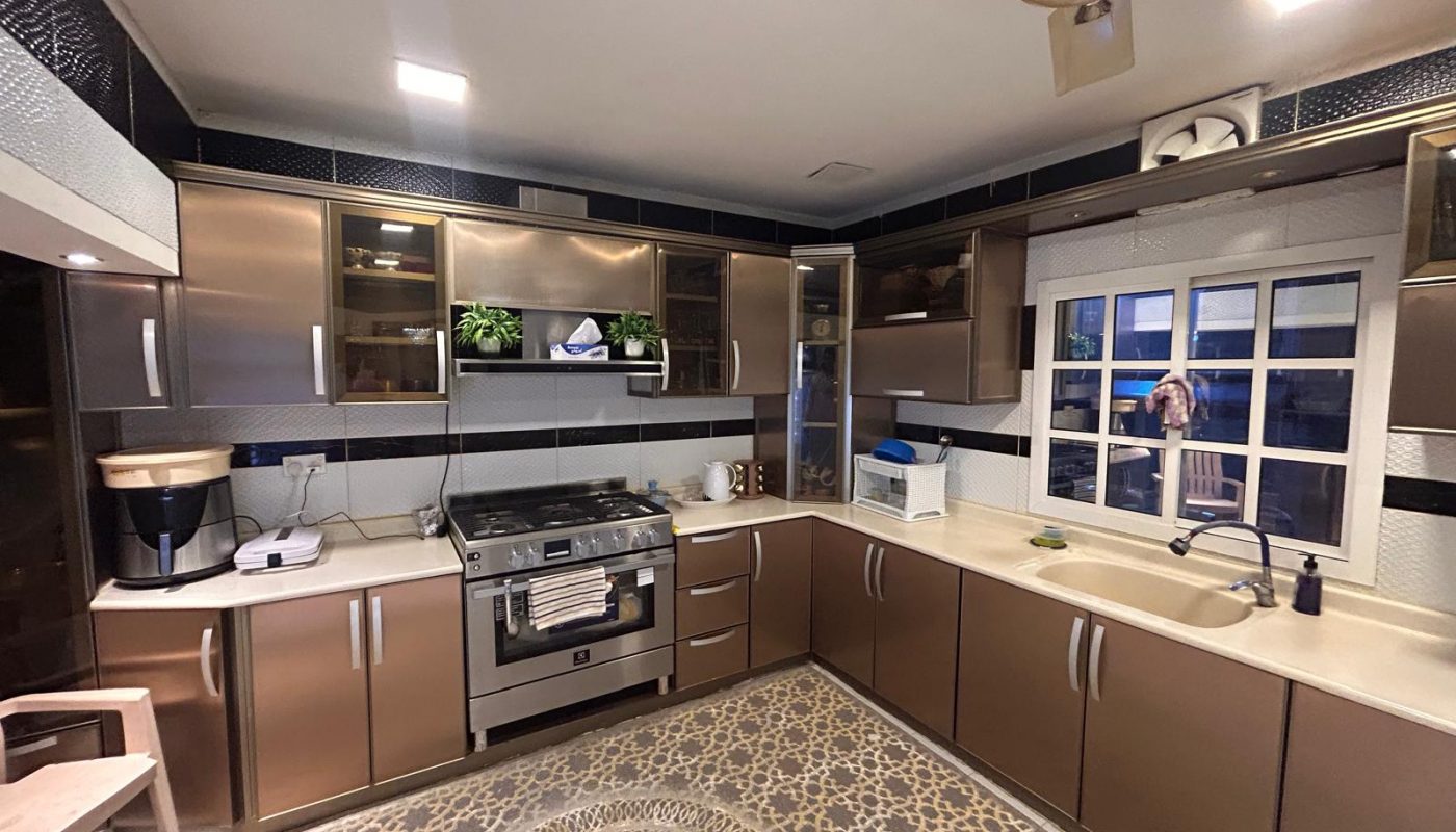 Modern kitchen with metallic brown cabinets, a gas stove, countertop appliances, white-tiled walls with black accents, a window above the sink, and a patterned gray floor rug.