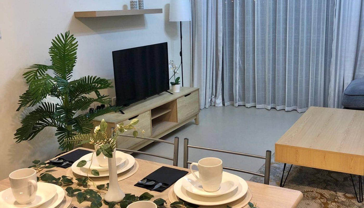 A dining table is set with white dishes and mugs in a modern living room of a 1-bedroom flat. The room features a TV on a wooden stand, a floor lamp, a plant, and a large window with sheer curtains. Located in Marassi Boulevard, this stylish flat is now for sale.