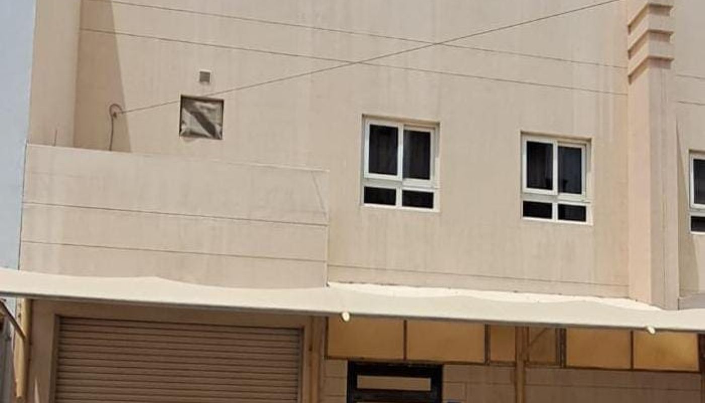 A beige two-story building with small square windows, a metal garage door, a black-striped entrance door, and a flat awning over the ground floor.