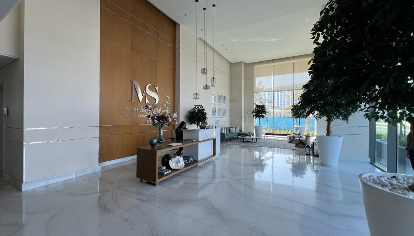 A modern office lobby with marble floors, large potted plants, a wooden reception desk, and "MS" logo on the wall behind the receptionist. Natural light enters through large windows.