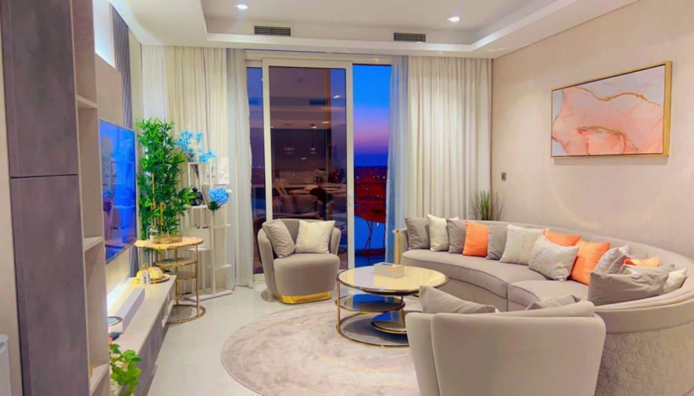 A modern living room with a curved sofa, armchairs, and a round coffee table. The room features a large window with curtains and a view of the city at dusk.