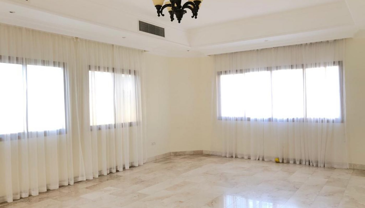 A spacious, empty room with marble flooring, large windows with sheer white curtains, a white ceiling, and a black chandelier.