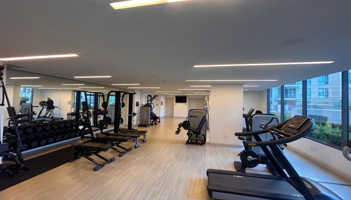 Modern gym with treadmills, weight racks, benches, and exercise machines on a light wood floor. Large mirrors and windows provide natural light.