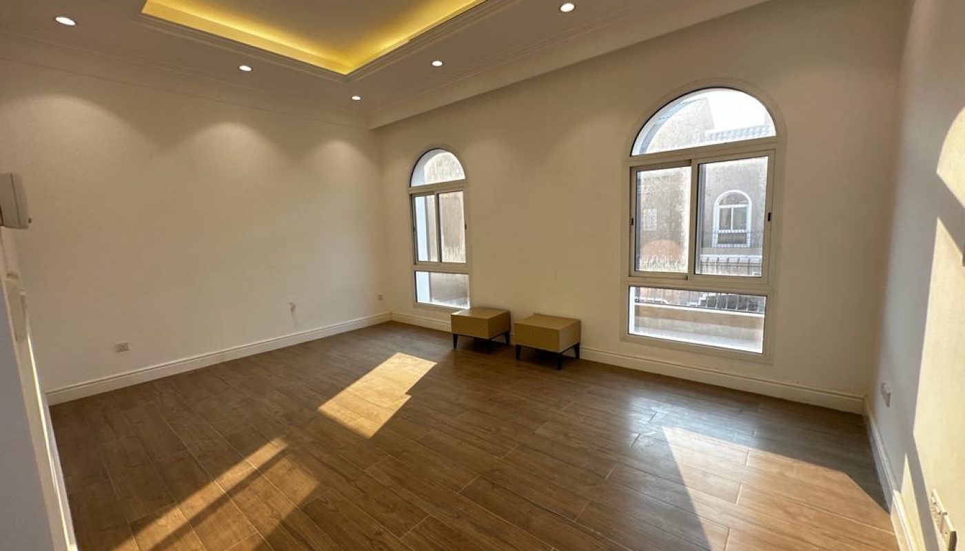 Sunlit empty room with wooden floor, recessed ceiling lights, two arched windows, and two small tables against the wall.