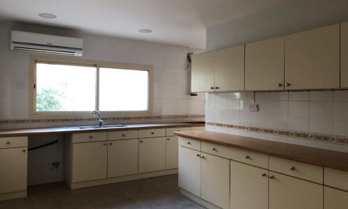 A clean, empty kitchen with white cabinets, beige countertops, a window, and an air conditioning unit above the sink.