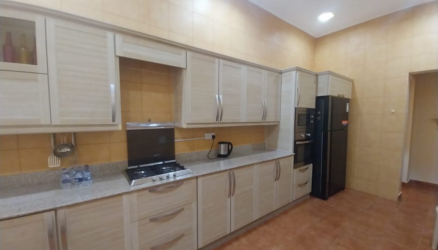 Modern kitchen with light wood cabinets, granite countertop, built-in stove, kettle, and black refrigerator against tan tiled walls and floor.