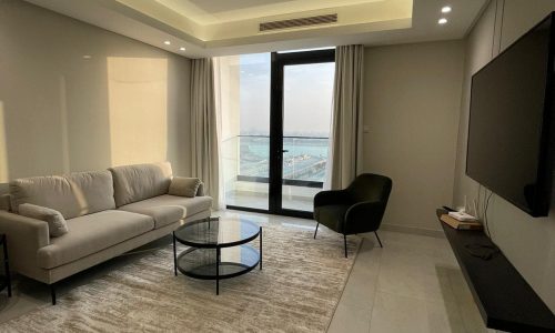 A modern, minimalist living room with a beige sofa, black armchair, glass coffee table, wall-mounted TV, and a large window overlooking a balcony with a water view. This AMWAJ flat for sale offers an exquisite blend of comfort and style.