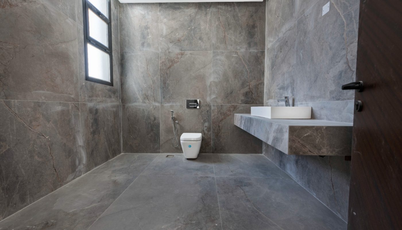 Spacious bathroom with gray stone tiles, a wall-mounted toilet, a bidet sprayer, a rectangular sink with a mirror, and two frosted windows.