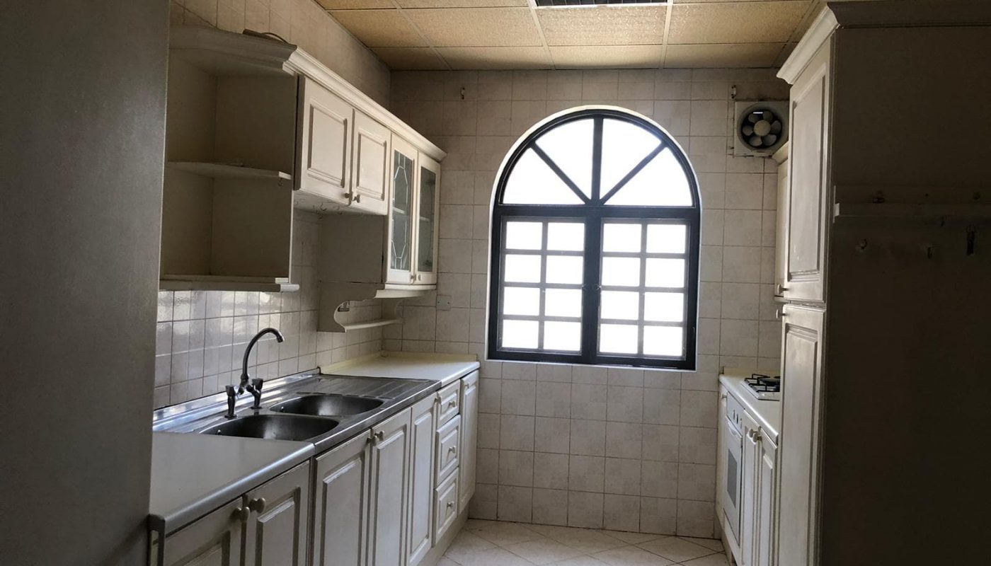 A kitchen with white cabinets, tiled walls, a double sink, gas stove, oven, and an arched window with a black frame, allowing natural light to fill the space.