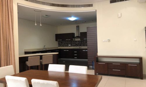 A modern open-plan kitchen and dining area featuring a wooden dining table, white chairs, a dark wood kitchen with bar stools, light-colored tiles, and recessed lighting.