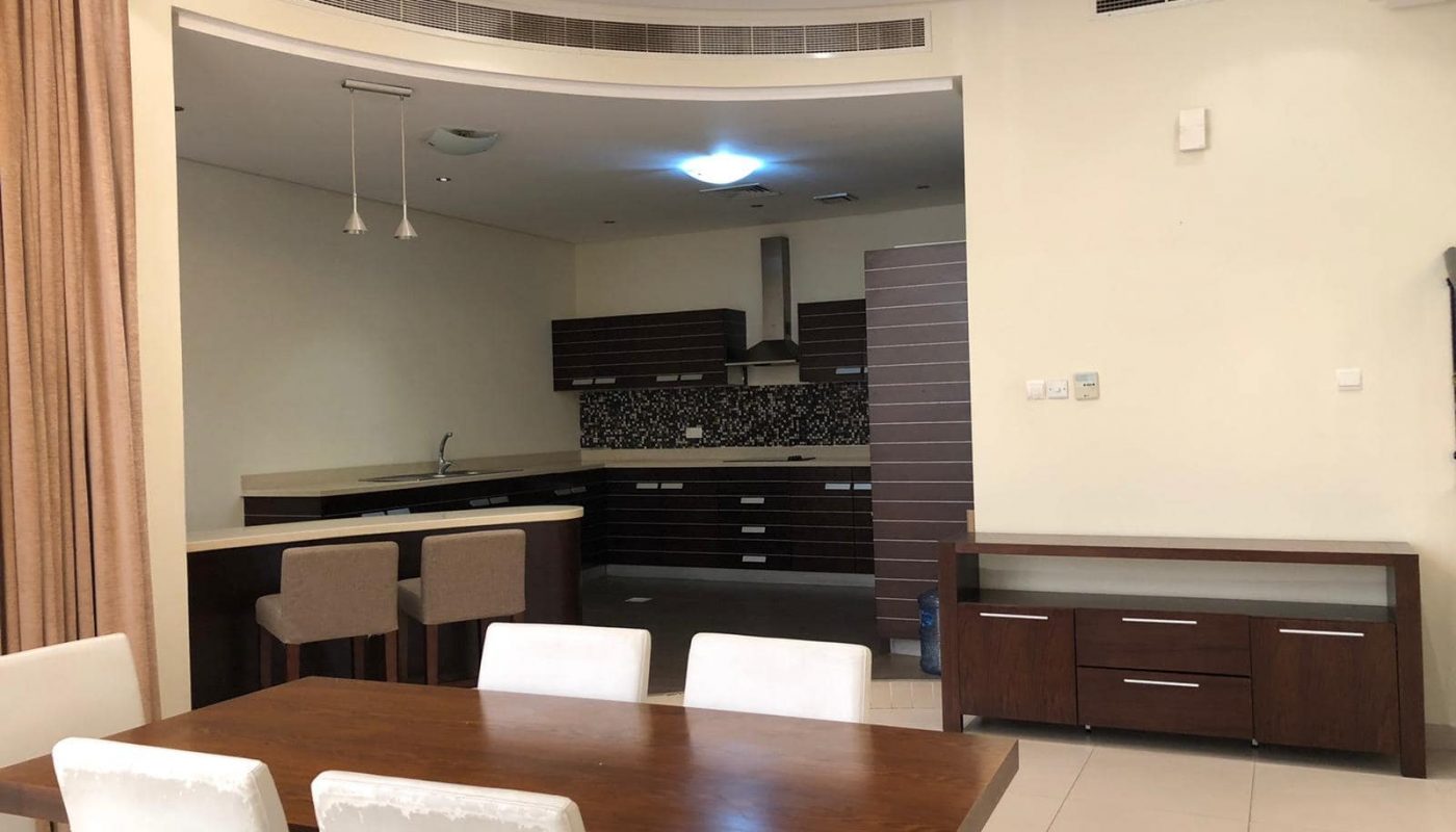 A modern open-plan kitchen and dining area featuring a wooden dining table, white chairs, a dark wood kitchen with bar stools, light-colored tiles, and recessed lighting.
