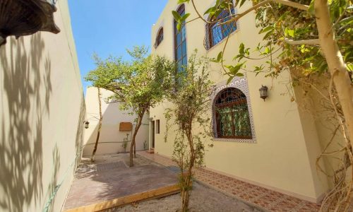 A shaded courtyard features a tree in the center, surrounded by beige buildings with large arched windows and decorative tiles. There are cobblestone paths and a wooden deck on the ground.