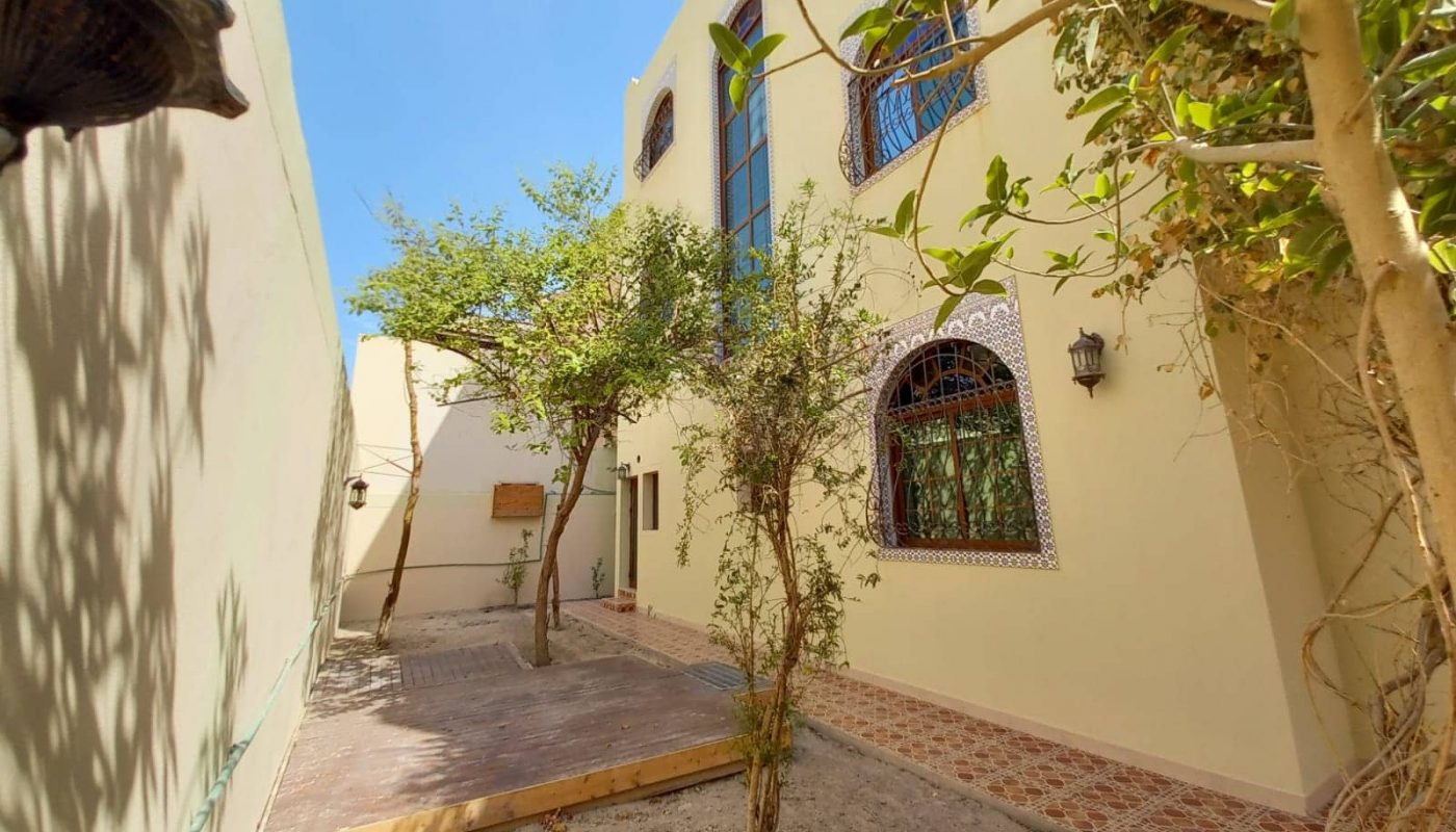 A shaded courtyard features a tree in the center, surrounded by beige buildings with large arched windows and decorative tiles. There are cobblestone paths and a wooden deck on the ground.