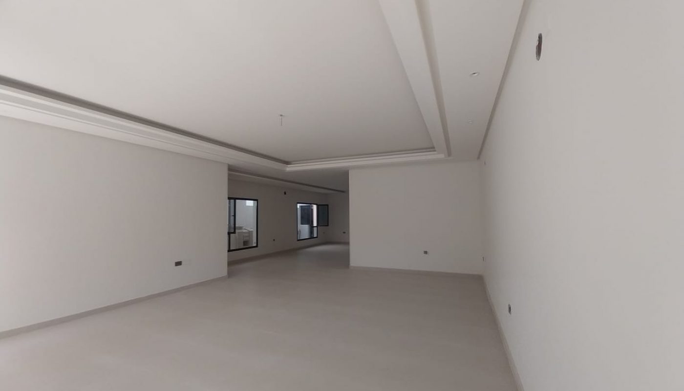 Empty, unfurnished, spacious white room with large windows, white tiled floor, and recessed ceiling lighting. Walls and ceiling are painted white, and there are no decorations or furniture.