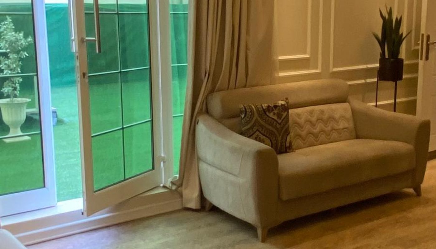 A beige sofa sits near a large glass door with cream curtains in a living room with wood flooring and a potted plant in the corner.