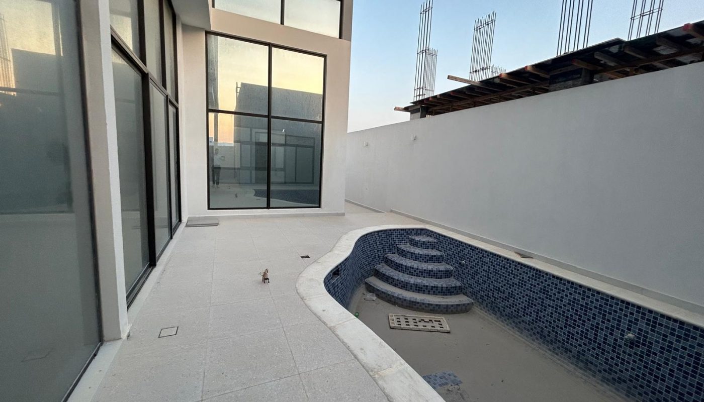 A modern house with large glass windows and an empty, tiled swimming pool in the backyard; unfinished construction is visible in the background.