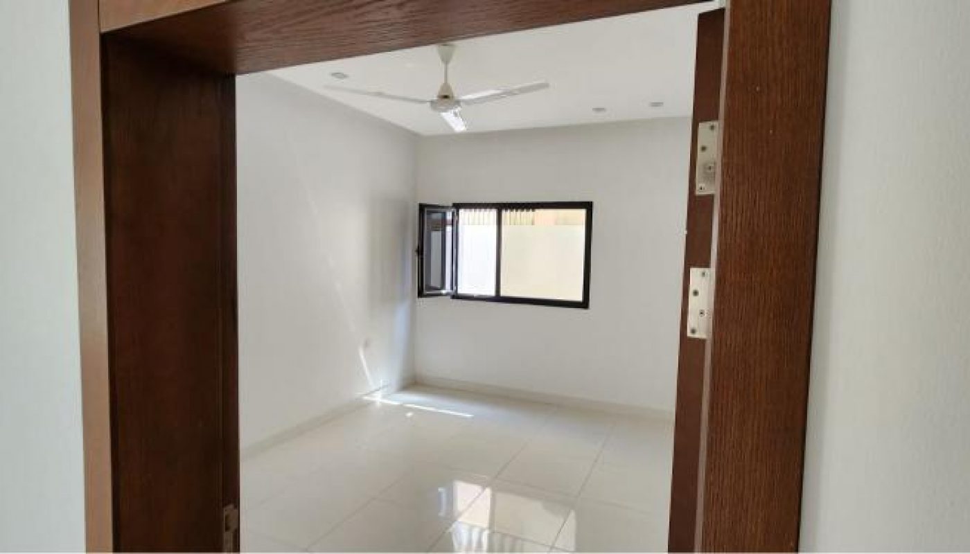 Empty, unfurnished room with white tiled floor, white walls, ceiling fan, and open window, viewed through a wooden door frame.