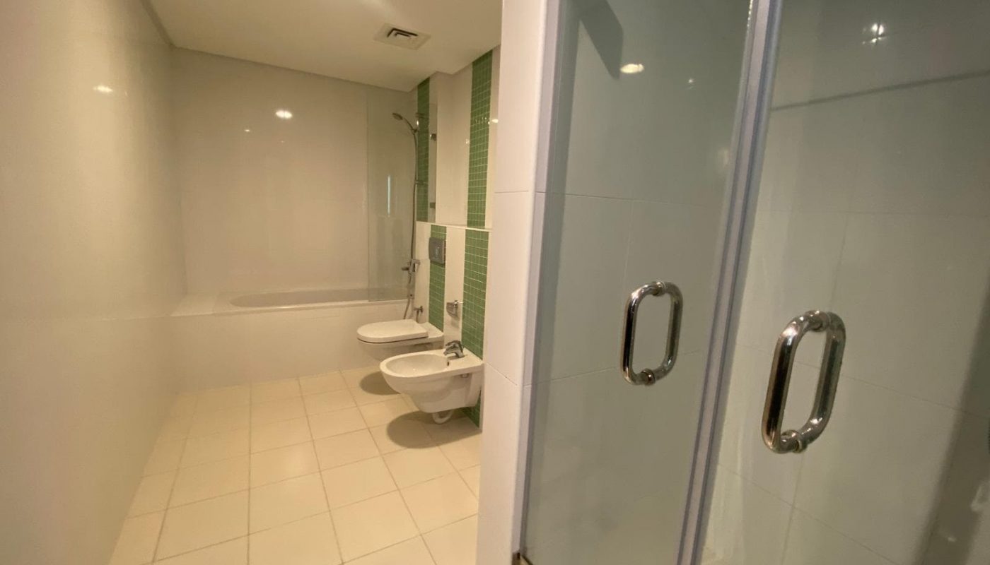 A spacious bathroom with a bathtub, toilet, bidet, and a glass-enclosed shower, featuring light-colored tiles and green accent wall by the fixtures.