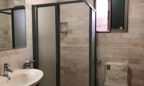 A bathroom with beige tiled walls, a walk-in shower with a glass door, a white sink with a wall-mounted faucet, a toilet, a small shelf, and a window with frosted glass.