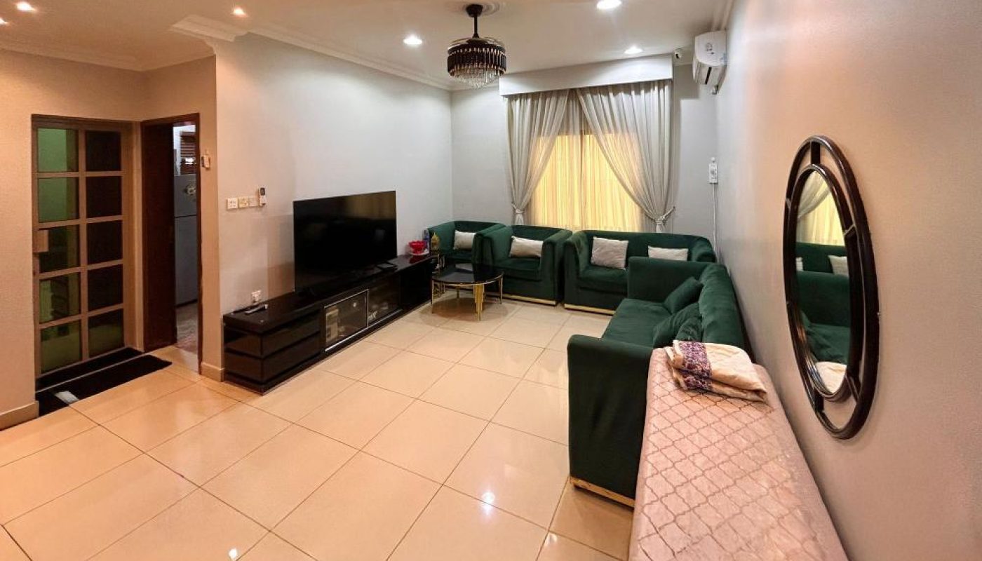 Spacious living room with tiled floor, green sectional sofa, TV on a stand, round mirror, chandelier, and large window with curtains.
