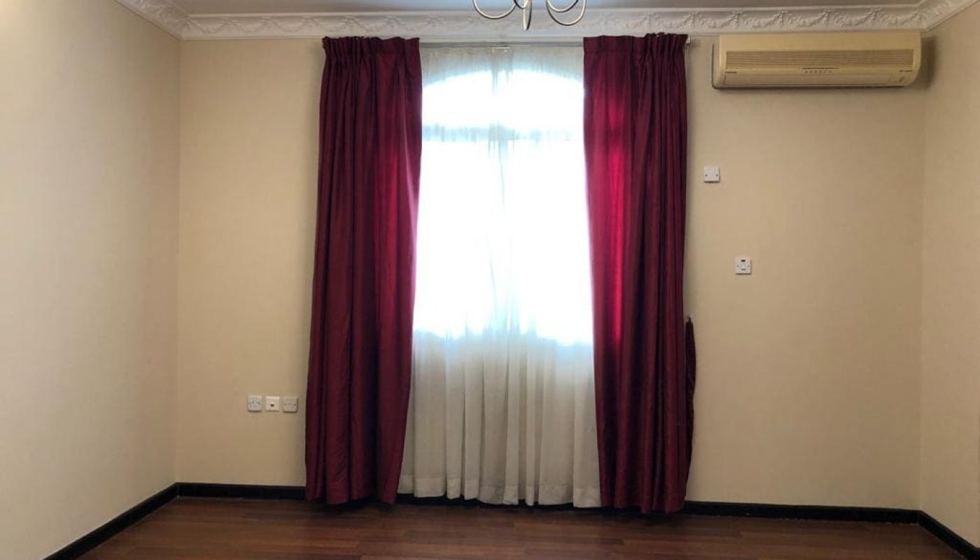 A semi-furnished room with beige walls, wooden flooring, a window adorned with red curtains, and an air conditioning unit mounted on the right wall. A chandelier adds elegance from the ceiling. This charming space is part of a 5-bedroom villa for rent.