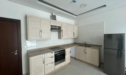 Modern kitchen in Al Hidd with beige cabinets, built-in oven, and a stainless steel refrigerator, featuring tiled walls and recessed ceiling lighting.