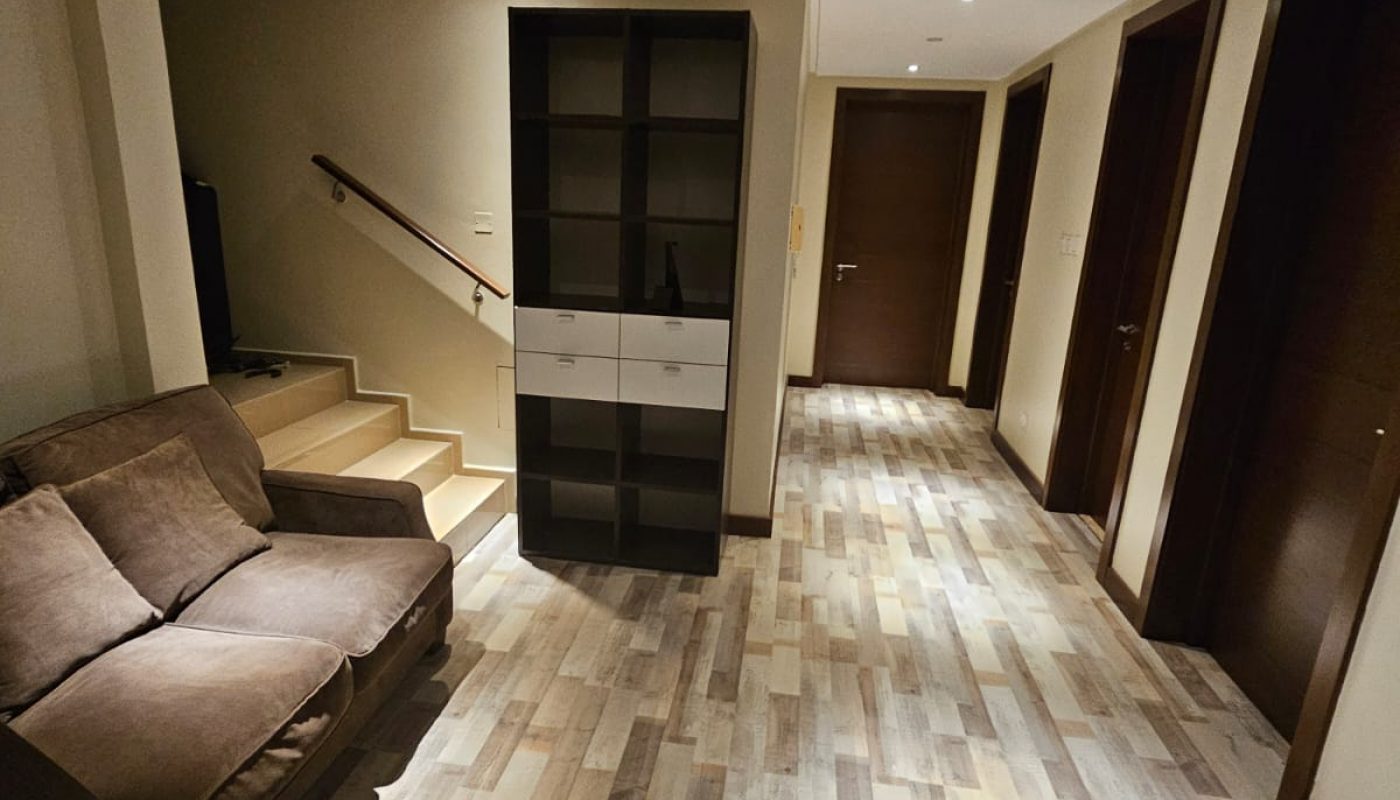 A hallway with wood-patterned flooring, a brown sofa, a black and white shelving unit, three closed doors, and a staircase leading upward. Ceiling lights illuminate the space.