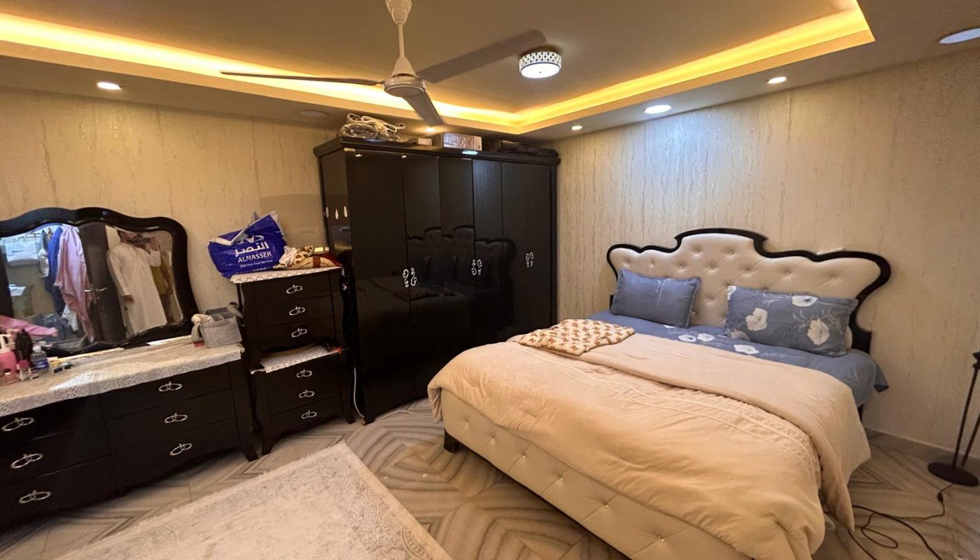 A bedroom with a double bed, two wardrobes, a dresser with a mirror, a ceiling fan, and warm recessed lighting. Various items are placed on the furniture.