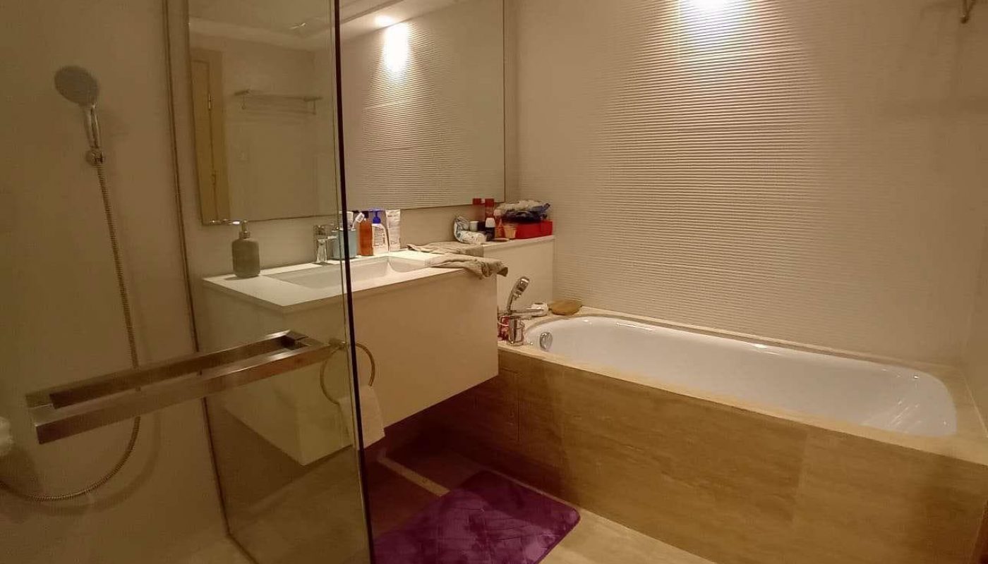 Modern bathroom with a glass shower, white sink, large mirror, bathtub with beige siding, purple bath mat, and various toiletries on the sink and tub ledge.