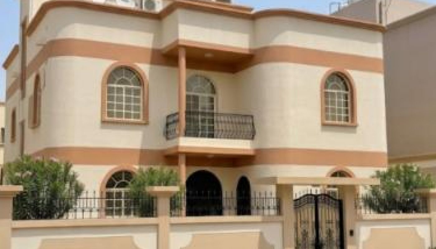 Two-story beige house with arched windows, small balcony, black gate, and a low wall, set on a paved street with desert landscaping in front.