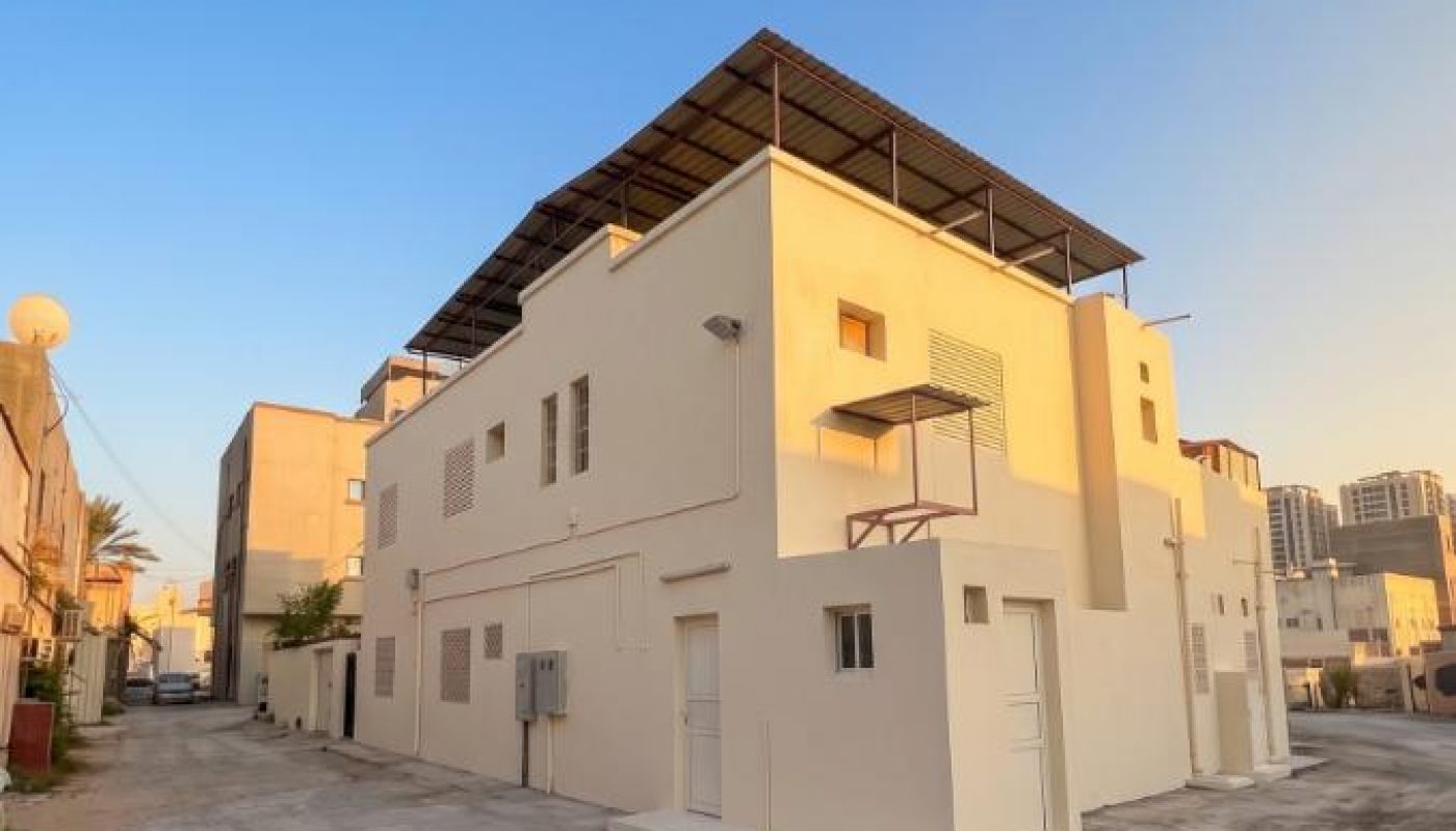 A beige two-story building with a rooftop structure stands on a quiet, sunlit street with a clear blue sky in the background.