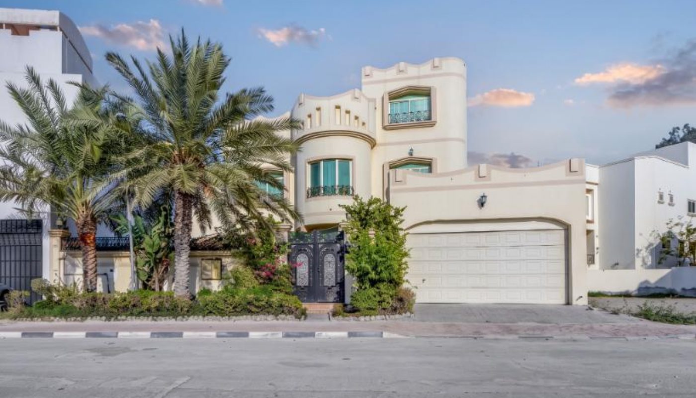 A cream-colored, two-story house with a double garage, arched windows, palm trees, and a gated entrance, viewed from across the street at sunset.