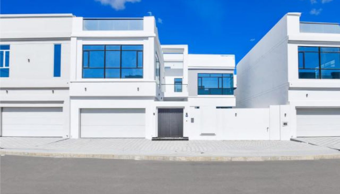 Modern white residential buildings with large blue-tinted windows, three garages, and a clear blue sky with scattered clouds above.
