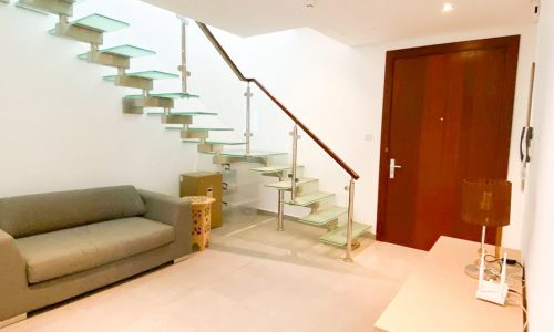 Discover a luxury apartment in the Amwaj area featuring a modern room with a glass staircase, a gray sofa, a wooden front door, and a beige console table with a lamp. The elegant space includes two wooden side tables and is now available for sale.