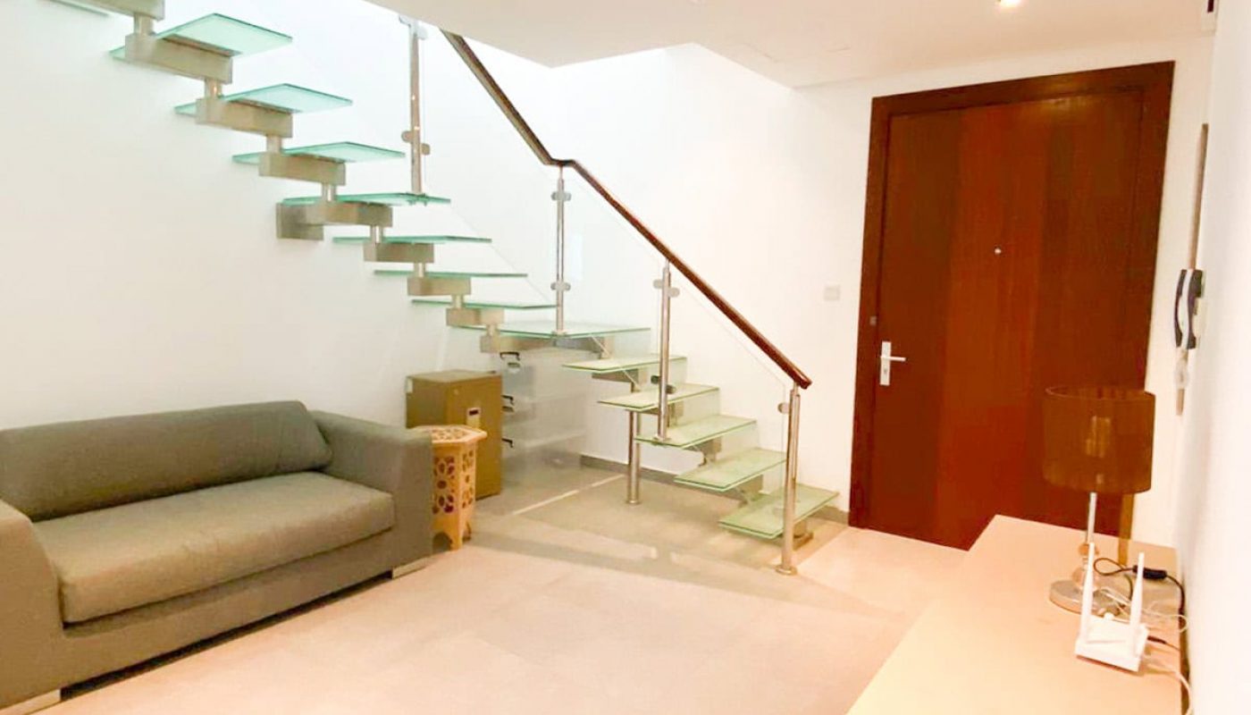 Discover a luxury apartment in the Amwaj area featuring a modern room with a glass staircase, a gray sofa, a wooden front door, and a beige console table with a lamp. The elegant space includes two wooden side tables and is now available for sale.