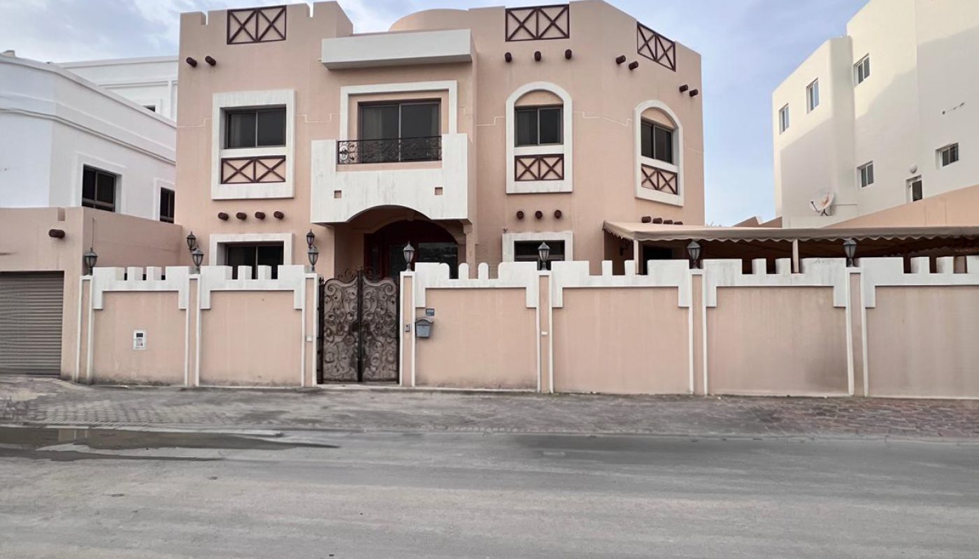 Two-story beige house with brown accents, decorative balcony railings, and a surrounding fence with a metal gate, situated on a paved street.