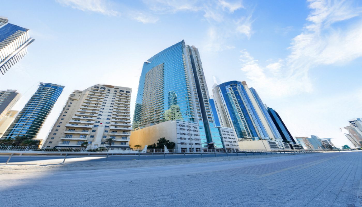 Tall modern skyscrapers line a wide, empty street under a clear blue sky, with sunlight reflecting off the glass buildings.