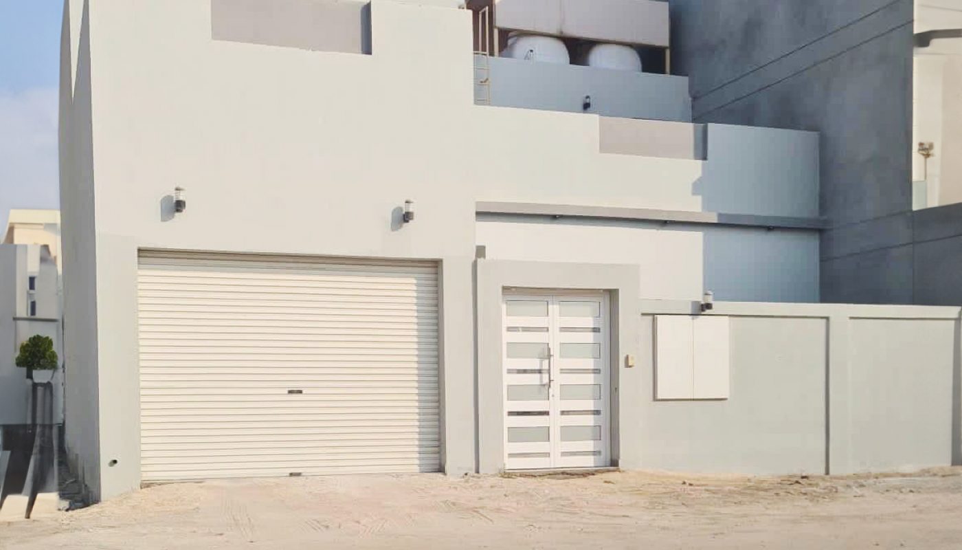 A modern, light gray building with a closed garage door, white gate, and cylindrical tanks on the roof, situated on a paved surface under a clear sky.