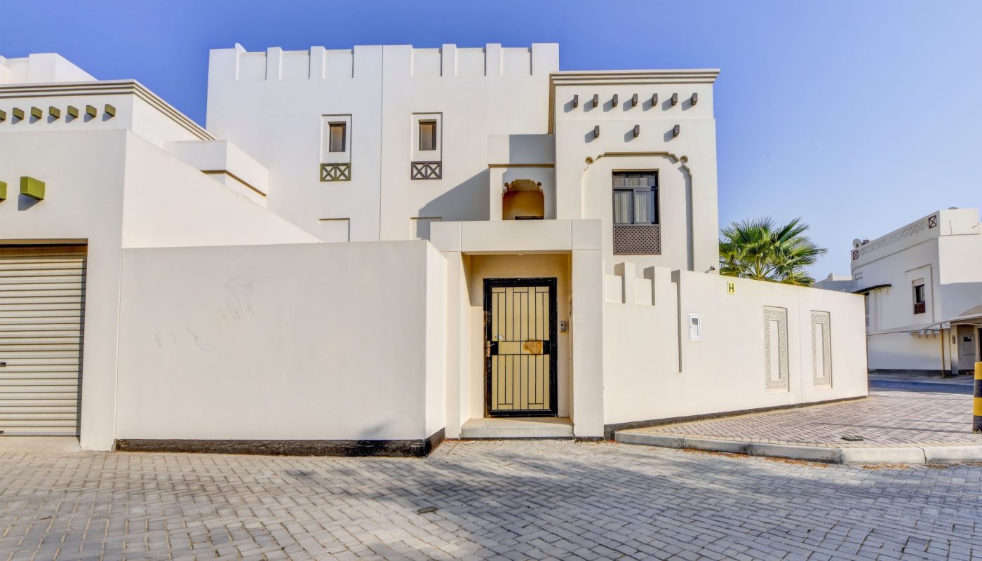 Modern white house with geometric design, flat roof, and decorative window grilles. Entry gate opens onto a paved driveway under a clear blue sky.