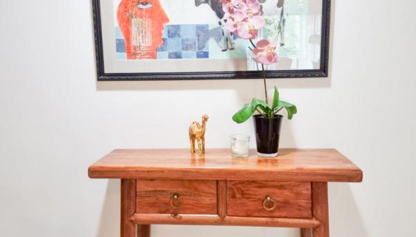 A wooden table with two drawers holds a potted orchid, a small gold camel figurine, and a candle. Above it hangs a colorful framed painting on a white wall.