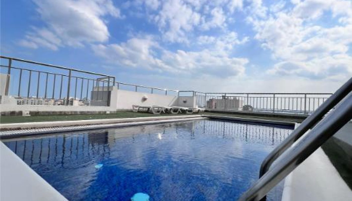 Rooftop swimming pool with blue water, surrounded by railing and lounge chairs, under a partly cloudy sky.