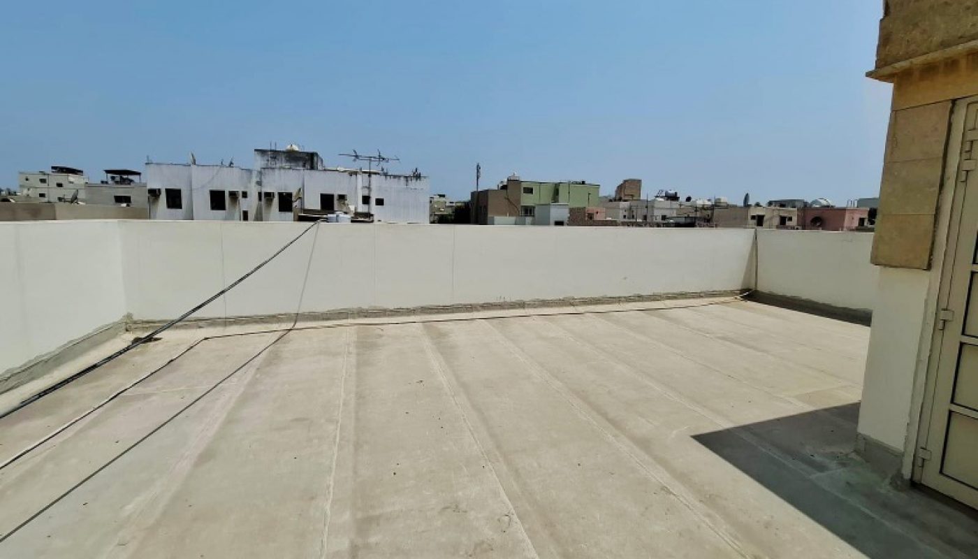 A flat, empty rooftop with concrete flooring under a clear blue sky, surrounded by white low walls and city buildings in the background.
