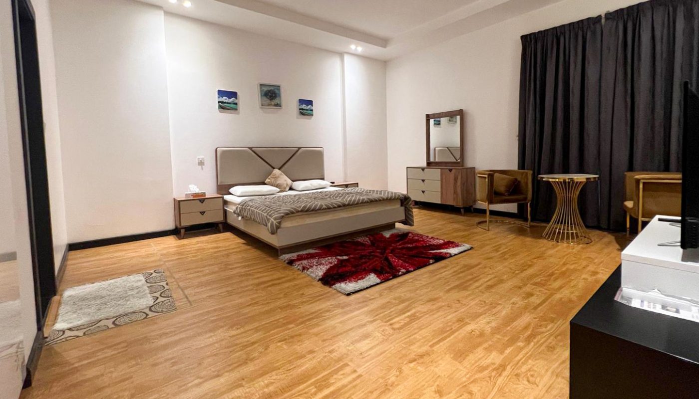 A spacious bedroom with a bed, bedside tables, a dresser with a mirror, a TV, dark curtains, wooden flooring, and simple wall art.