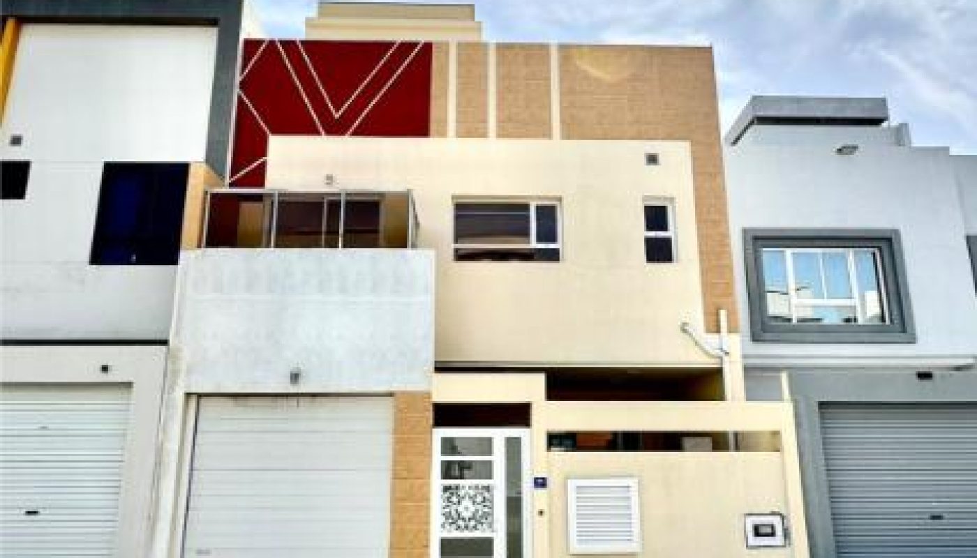 Three-story modern townhouse with a beige and red facade, a garage door, and a glass main entrance, under a partly cloudy sky.