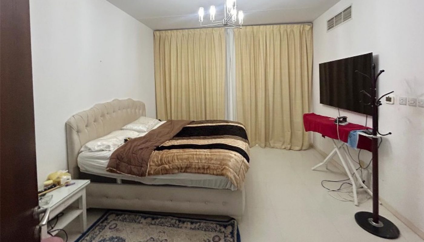 A bedroom with a bed, nightstand, rug, wall-mounted TV, TV stand, and beige curtains covering a window or door.