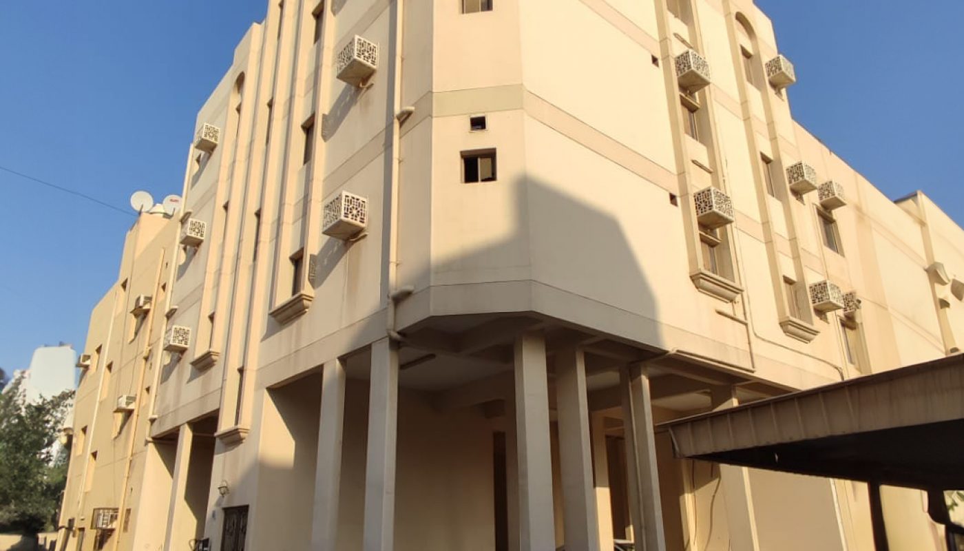 A beige three-story apartment building with rectangular columns, small balconies, and a covered parking area, photographed from a low angle on a clear day.